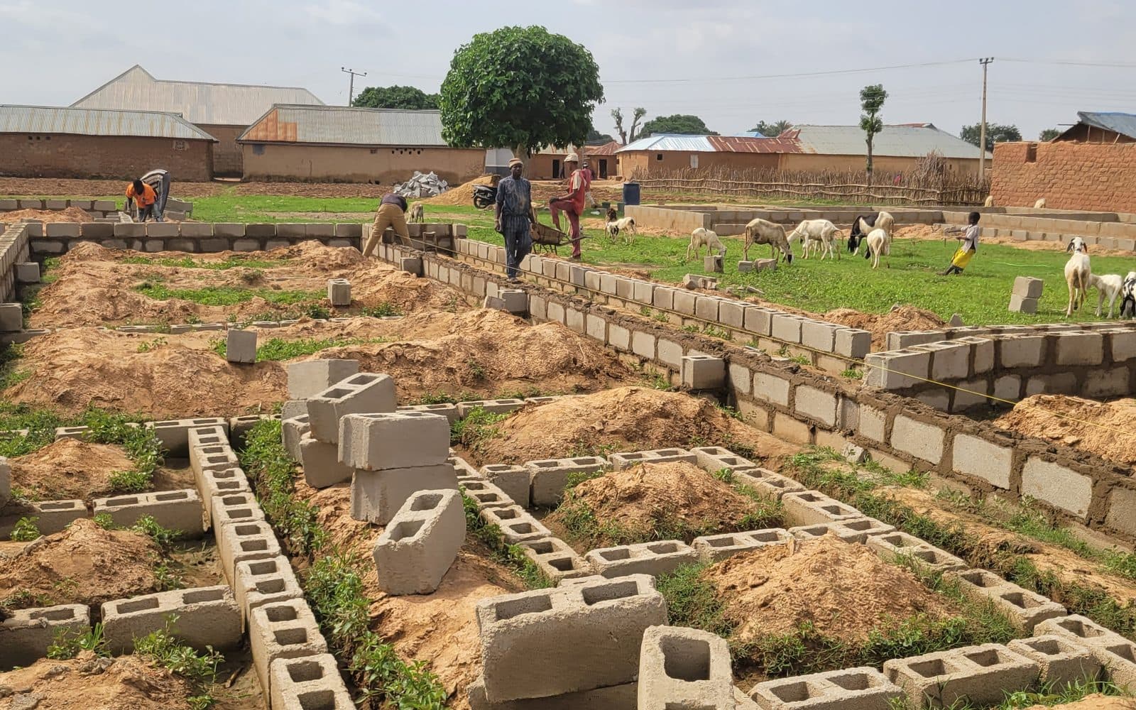 Block foundation walls laid out across the Katanga build site, with workers running a string line and the village beyond