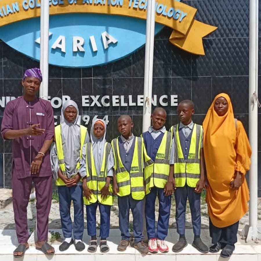 Pupils in reflective vests with two staff members at the Nigerian College of Aviation Technology gate in Zaria
